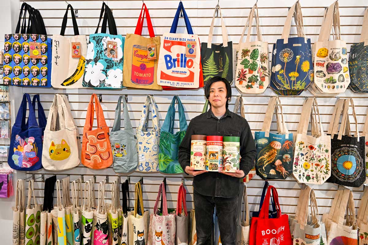 店長の久保さん、プラスチックを使用しないエコなパズルを手に、カラフルなデザインのエコバッグの前で。GIFTS AND THINGS。Photo by Koichi Saito/Japan Canada Today